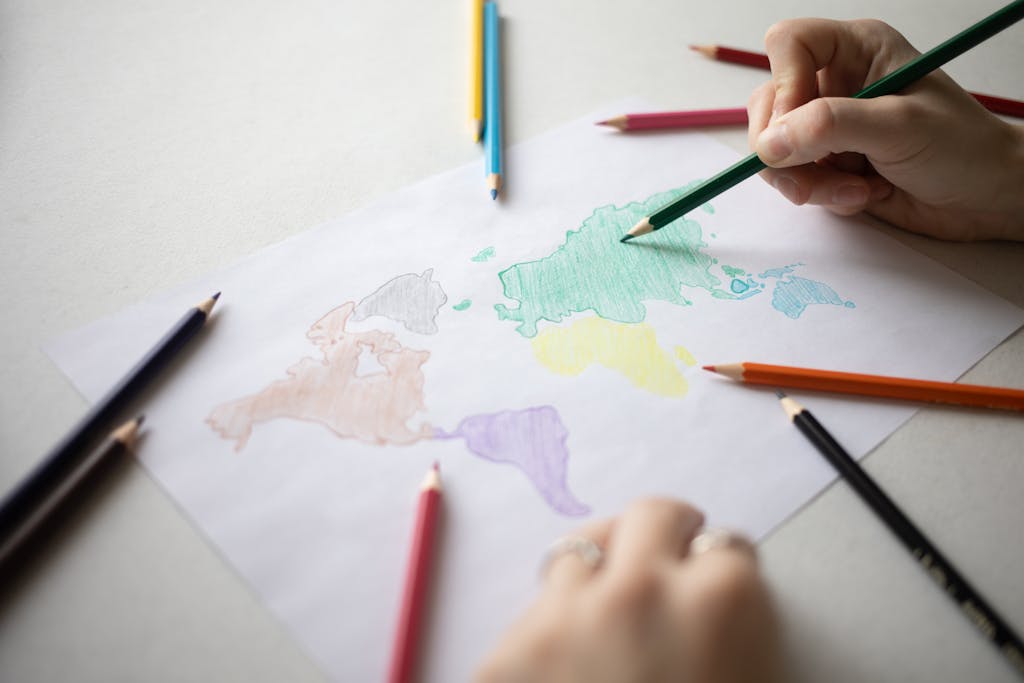 Crop anonymous person with colored pencils coloring continents on white paper on table with various scattered stationery in light room